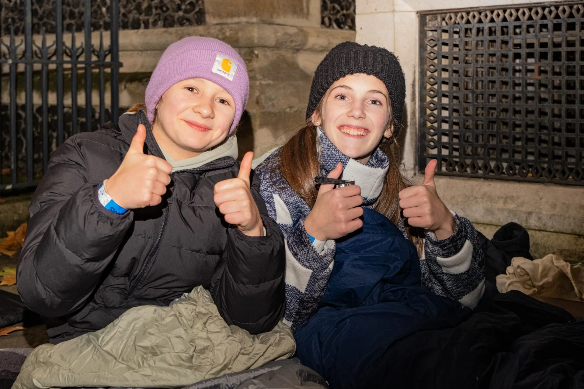 Two girls wearing coats and sleeping bags smiling and thumbs up at the camera