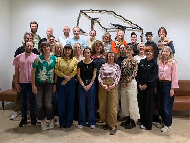 A large group of twenty-three people posing for a photo in front of a wall-mounted wire sculpture of a mountain range.